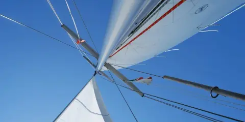 Vue plongeante sur le mât et les voiles d'un voilier, le ciel bleu intense en arrière-plan. L'image capture l'essence de la navigation, le vent gonflant les voiles.