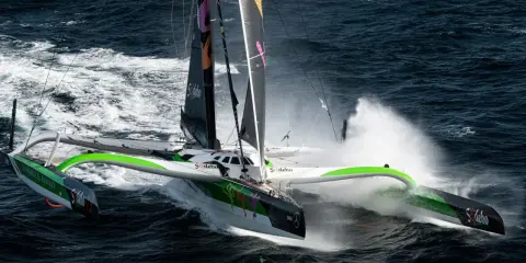 Le trimaran Sodebo, un bateau ultime, fend les vagues sombres, projetant des gerbes d'eau dans une course effrénée.