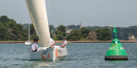 Voile blanche sur l'eau, le voilier "Bihan 6.50" navigue avec deux personnes à bord. Une bouée verte marque le passage.