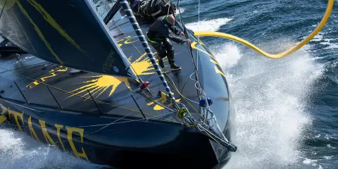 Un skipper manœuvre sur l'IMOCA Bureau Vallée, le bateau noir et jaune fend les vagues dans un embrun spectaculaire.