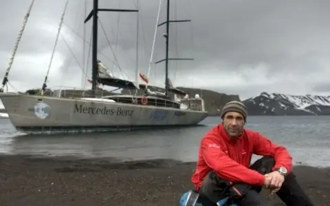 Mike Horn, l'explorateur, pose près de son bateau "Mercedes-Benz" sur une plage rocheuse, avec des montagnes enneigées en arrière-plan.