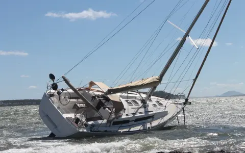 Un voilier échoué, incliné sur le côté, témoigne d'une possible collision bateau. Les vagues s'écrasent sur sa coque blanche sous un ciel bleu.