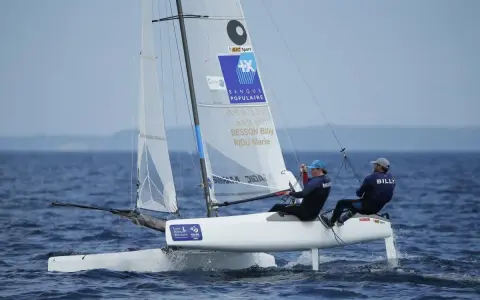 Deux marins, dont Billy Besson, naviguent sur un catamaran blanc par temps calme.