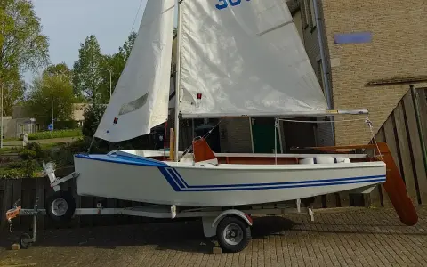 Un petit voilier blanc et bleu, un vaurien bateau prêt pour l'aventure, est sur sa remorque.
