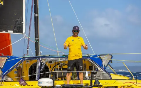 Arnaud Boissières, souriant, fait un double pouce levé sur son voilier jaune et bleu, prêt pour une nouvelle aventure en mer.