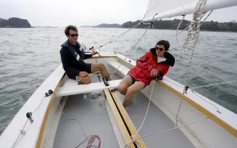 Deux personnes profitent d'une journée ensoleillée sur un voilier dayboat, naviguant sur une mer calme.