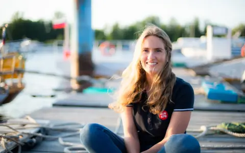 Irina Gracheva, souriante, assise sur un ponton en bois, avec des bateaux et des bouées en arrière-plan.