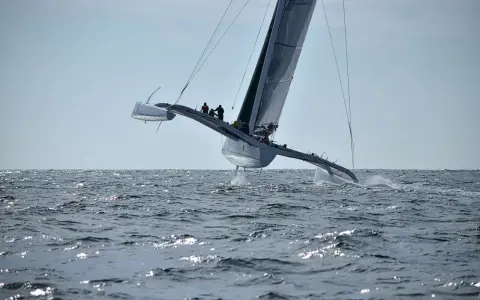 Un maxi trimaran fend les vagues, ses coques latérales hors de l'eau, propulsé par une voile immense. L'équipage est visible sur le pont.