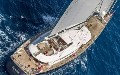Le yacht "Bayesian" navigue sur des eaux bleues, ses voiles blanches gonflées par le vent. L'équipage en rouge s'affaire sur le pont en bois.