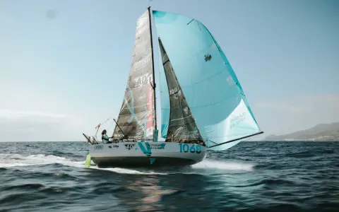 Un voilier de course, le numéro 1068, navigue sur une mer agitée. Les voiles, l'une noire et l'autre bleu clair, portent le nom "la maison perchée". Thais Le Cam est à la barre.