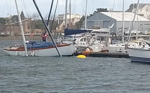 Un voilier blanc et bois, bâché, est partiellement immergé dans un port. Un homme en gilet rouge est à bord.
