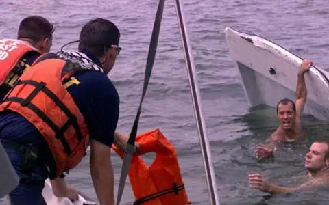 Sauvetage après un naufrage de bateau. Des secouristes en gilets orange aident des hommes dans l'eau, près d'une embarcation renversée.