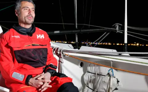 Giancarlo Pedote, skipper en tenue de voile rouge, sur un bateau la nuit, avec des lumières de ville en arrière-plan.