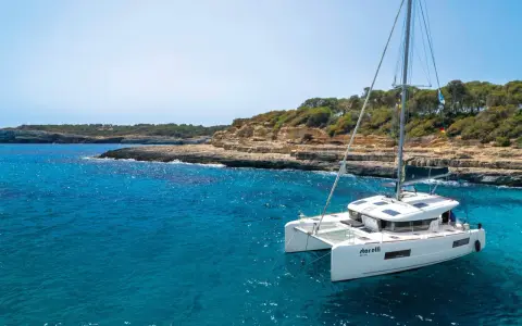 Un catamaran blanc, le "StarElli", navigue dans des eaux turquoise près d'une côte rocheuse et verdoyante. Ce safran catamaran offre une vue idyllique.