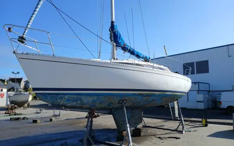 Voilier blanc sur bers, coque bleue usée, sous un ciel clair. Le mât est dressé, la grand-voile bleue enroulée. Premier 29 avis.
