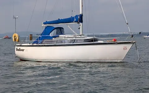 Voilier blanc Dufour 2800 à l'ancre, avec une bâche bleue sur la cabine. Un remorqueur orange est visible au loin.