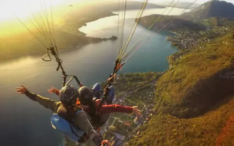 Vol en parapente biplace au-dessus d'un lac, avec un paysage de montagne et de village. Philippe Jeantot, malgré un accident passé, continue de voler.