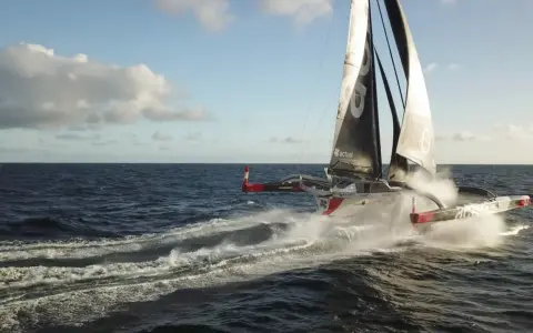 Le trimaran "Actual" d'Yves Le Blevec fend les vagues, propulsé par le vent sous un ciel bleu.