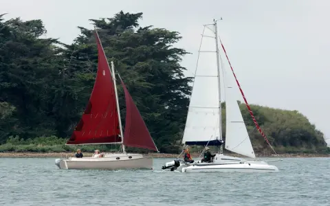 Deux voiliers naviguent sur l'eau, l'un avec une voile rouge, l'autre avec une voile blanche. On dirait un **bateau corsaire** prêt pour l'aventure.