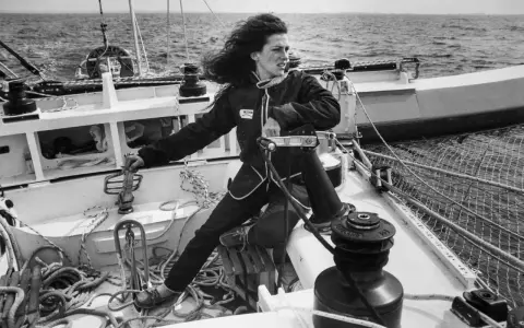 Une jeune femme aux cheveux longs, vêtue d'une combinaison de marin, manœuvre un winch sur un voilier. L'image évoque l'esprit d'aventure de Jacques Arthaud.