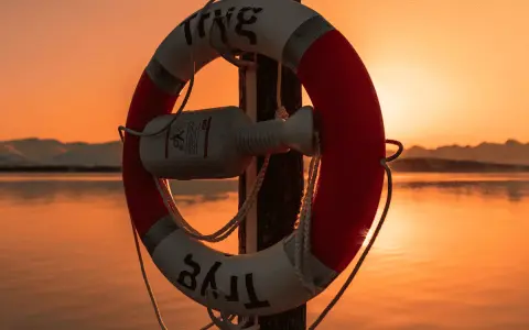 Bouée de sauvetage rouge et blanche sur un poteau, au bord d'une voie d'eau calme, avec un bateau à l'horizon au coucher du soleil.