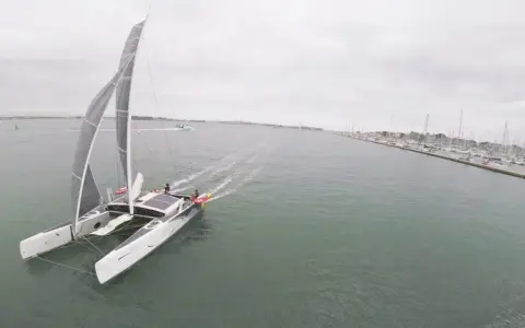 Un catamaran TS3 navigue sur une mer calme, avec un port rempli de voiliers à droite.