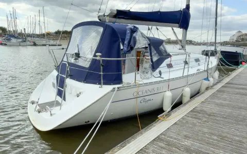 Un voilier blanc, un Océanis 311 Clipper, est amarré à un quai en bois. Des pare-battages blancs protègent sa coque.