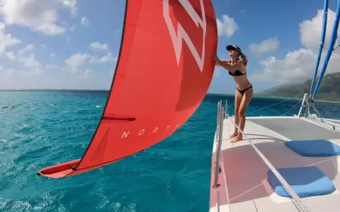 Une femme en bikini ajuste la voile rouge d'un petit catamaran habitable. L'eau turquoise et le ciel bleu créent un décor paradisiaque.