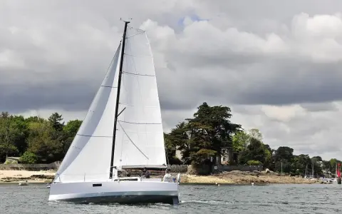 Voilier blanc moderne naviguant sur l'eau, avec des avis sur le Pogo 30. Paysage côtier verdoyant en arrière-plan.