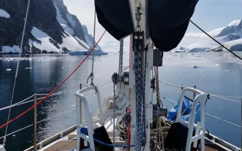 Voile sur un voilier dans un fjord glacé, avec des montagnes enneigées et des icebergs. Le **pen duick 600** navigue paisiblement.