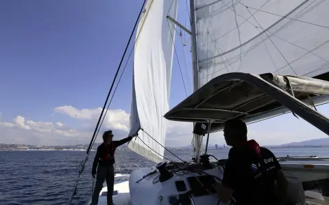 Deux hommes sur un catamaran, l'un ajuste la voile pour se mettre à la cape, tandis que l'autre tient la barre. Le soleil brille sur la mer bleue.