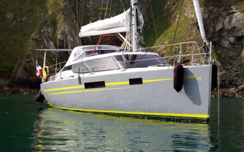 Le voilier "Bi-loup 109" gris avec des bandes jaunes, ancré près d'une falaise verdoyante.