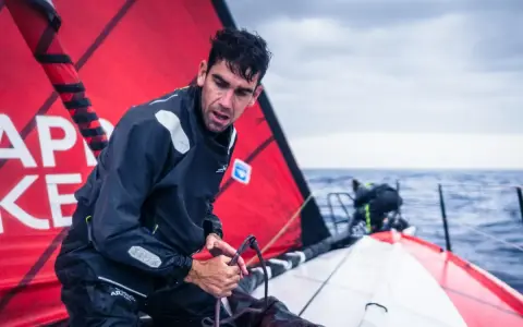 Gaston Morvan, marin concentré, ajuste une corde sur un voilier par temps couvert. La voile rouge porte des inscriptions blanches.
