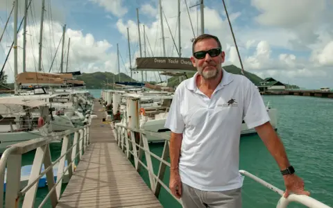 Loïc Bonnet, un homme souriant, pose sur un ponton bordé de voiliers. Il porte un polo Dream Yacht Charter, prêt pour une aventure en mer.
