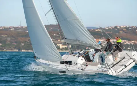 Un voilier First 27.7 navigue sur une mer agitée, avec quatre personnes à bord. Le soleil brille sur les voiles blanches et le paysage côtier en arrière-plan.