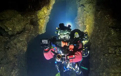 Plongée dans une grotte sous-marine, Xavier Meniscus filme les parois rocheuses éclairées par ses lampes.
