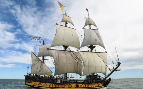 Le Shtandart, bateau historique aux voiles blanches, navigue sur une mer agitée sous un ciel nuageux.