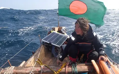 Capucine Trochet navigue sur une mer agitée, le drapeau du Bangladesh flottant au vent. Un panneau solaire est visible sur le bateau.