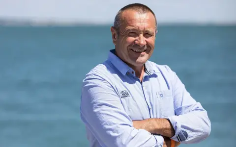 Lalou Roucayrol, souriant, bras croisés, devant la mer. Il porte une chemise bleue rayée avec un logo sur la poitrine.