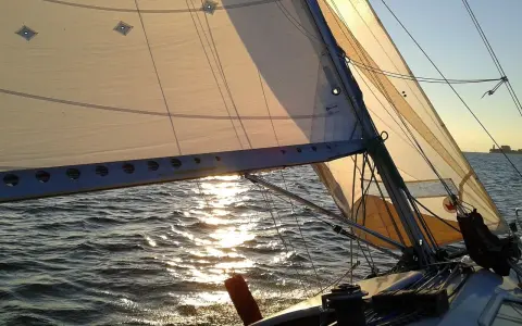 Voile d'un bateau au coucher du soleil, reflets dorés sur l'eau. La balancine du bateau est bien tendue.