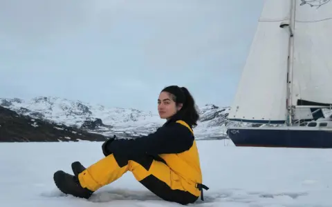 Tamara Klink, en tenue de ski jaune et noire, assise sur la neige, avec un voilier et des montagnes enneigées en arrière-plan.