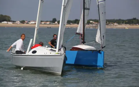 Deux voiliers naviguent côte à côte. Sur le premier, un homme en blanc manœuvre. Le second, d'un bleu vif, est un aloes 18.
