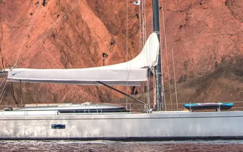 Un voilier Swan 76, avec une voile blanche déployée, navigue près d'une falaise rouge. Un kayak est rangé sur le pont.