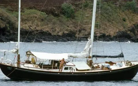 Le voilier Askoy 2, avec un homme sur le pont, navigue près d'une côte rocheuse et verdoyante.