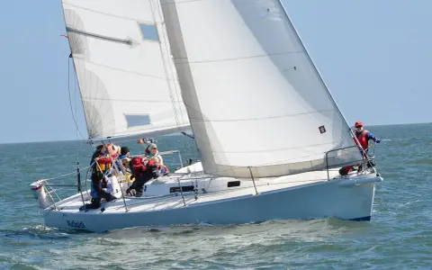 Un voilier blanc navigue sur une mer agitée, avec plusieurs personnes à bord. Le soleil brille, les voiles sont gonflées par le vent.