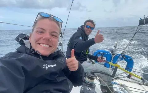 Hugo Dhallenne et sa coéquipière souriants sur un voilier, pouces levés, en pleine mer sous un ciel couvert.