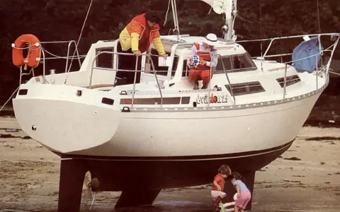 Le voilier "Évasion 29" échoué sur la plage, avec deux enfants jouant à ses pieds et deux adultes à bord.