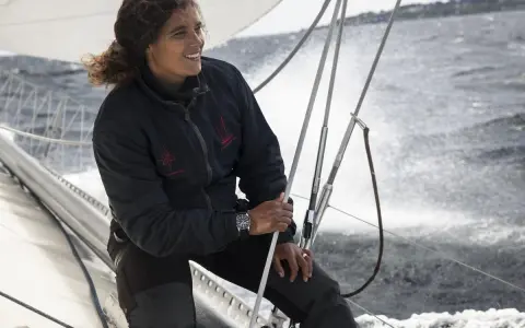 Marie Tabarly, souriante, tient la barre d'un voilier, le vent dans ses cheveux, naviguant sur une mer agitée.