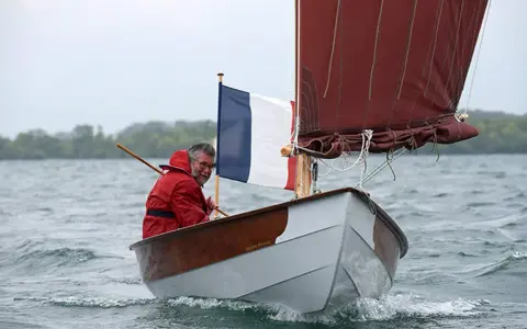 Un homme en ciré rouge navigue sur son bateau, fier du résultat de son travail pour construire son bateau. Le drapeau français flotte fièrement.