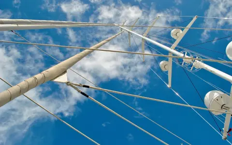 Vue plongeante sur le gréement d'un bateau, mâts blancs et cordages fins se découpant sur un ciel bleu et nuageux.